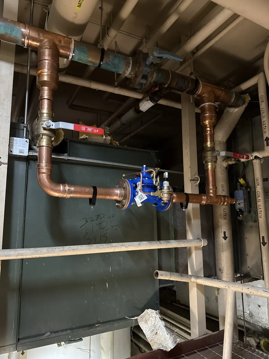 Large-diameter copper piping with a blue valve assembly running along the ceiling of a mechanical room above a dark green boiler.
