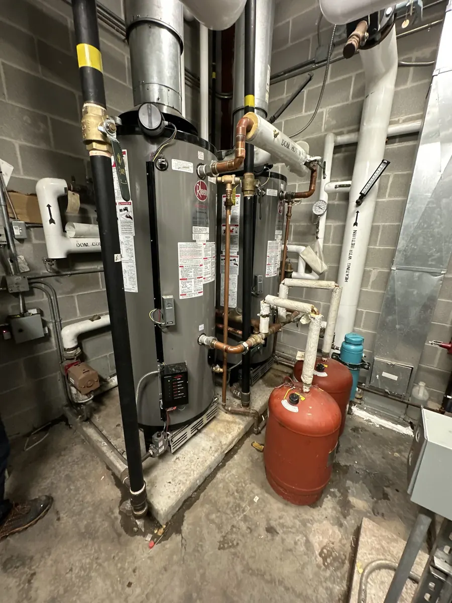 A tall water heater beside black gas piping with yellow tags, red expansion tanks and white vent pipes in a basement.